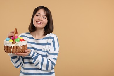 Easter celebration. Happy woman with wicker basket full of painted eggs on beige background, space for text