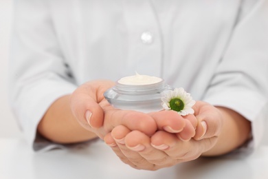 Photo of Female dermatologist holding jar of skin care product at table, closeup