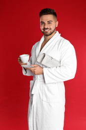 Photo of Young man in bathrobe with cup of coffee and newspaper on red background