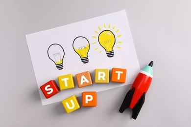 Photo of Colorful cubes with words Start Up, toy rocket and drawn lamps on grey background, flat lay