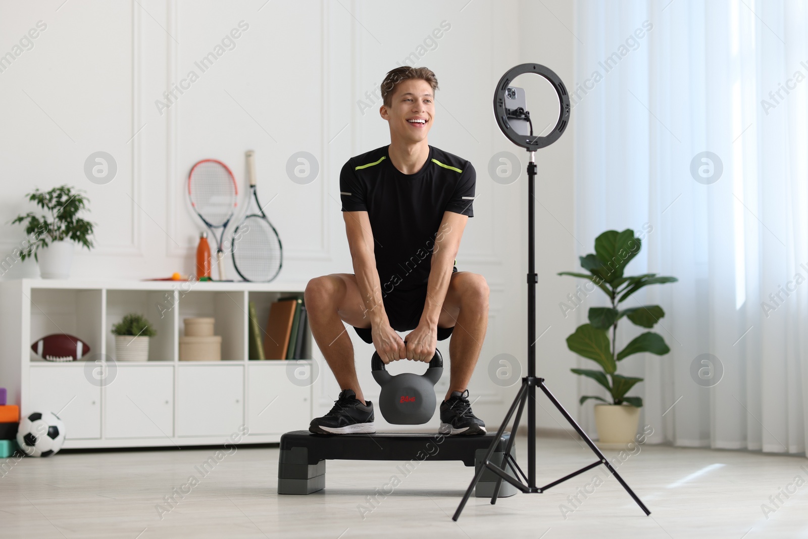 Photo of Smiling sports blogger training with kettlebell while streaming online fitness lesson at home