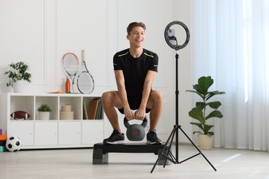 Photo of Smiling sports blogger training with kettlebell while streaming online fitness lesson at home