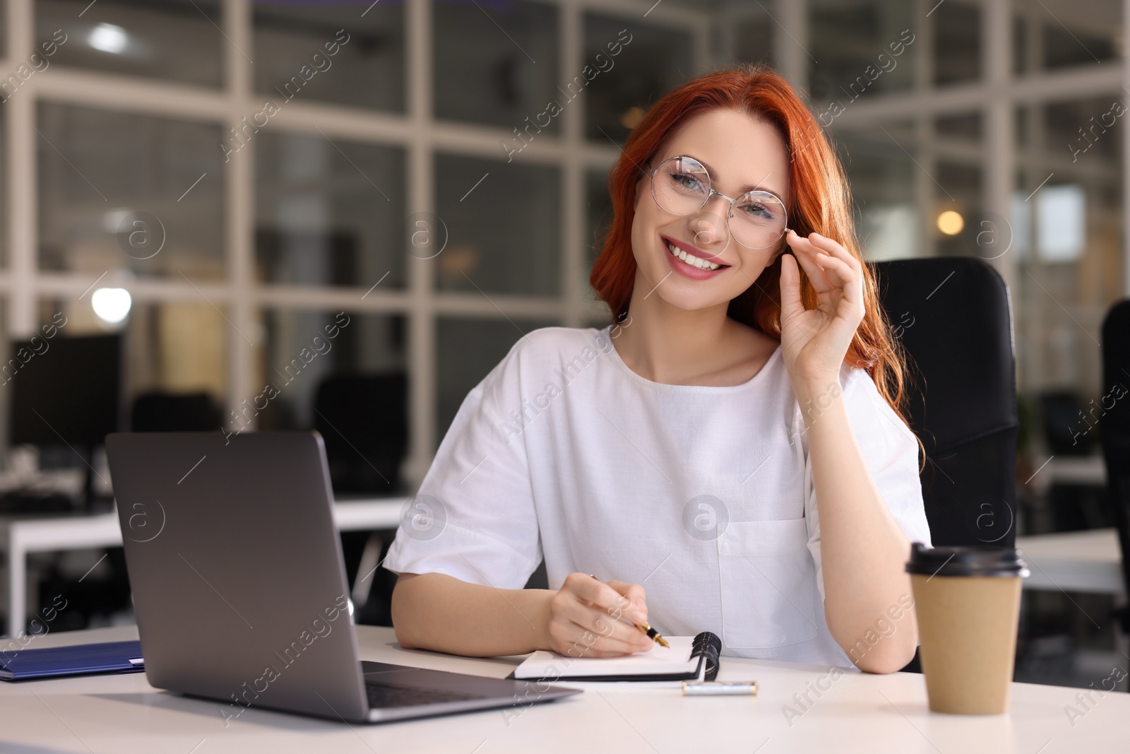 Photo of Happy woman taking notes while working with laptop at white desk in office