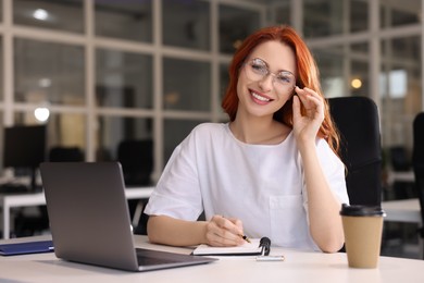 Photo of Happy woman taking notes while working with laptop at white desk in office