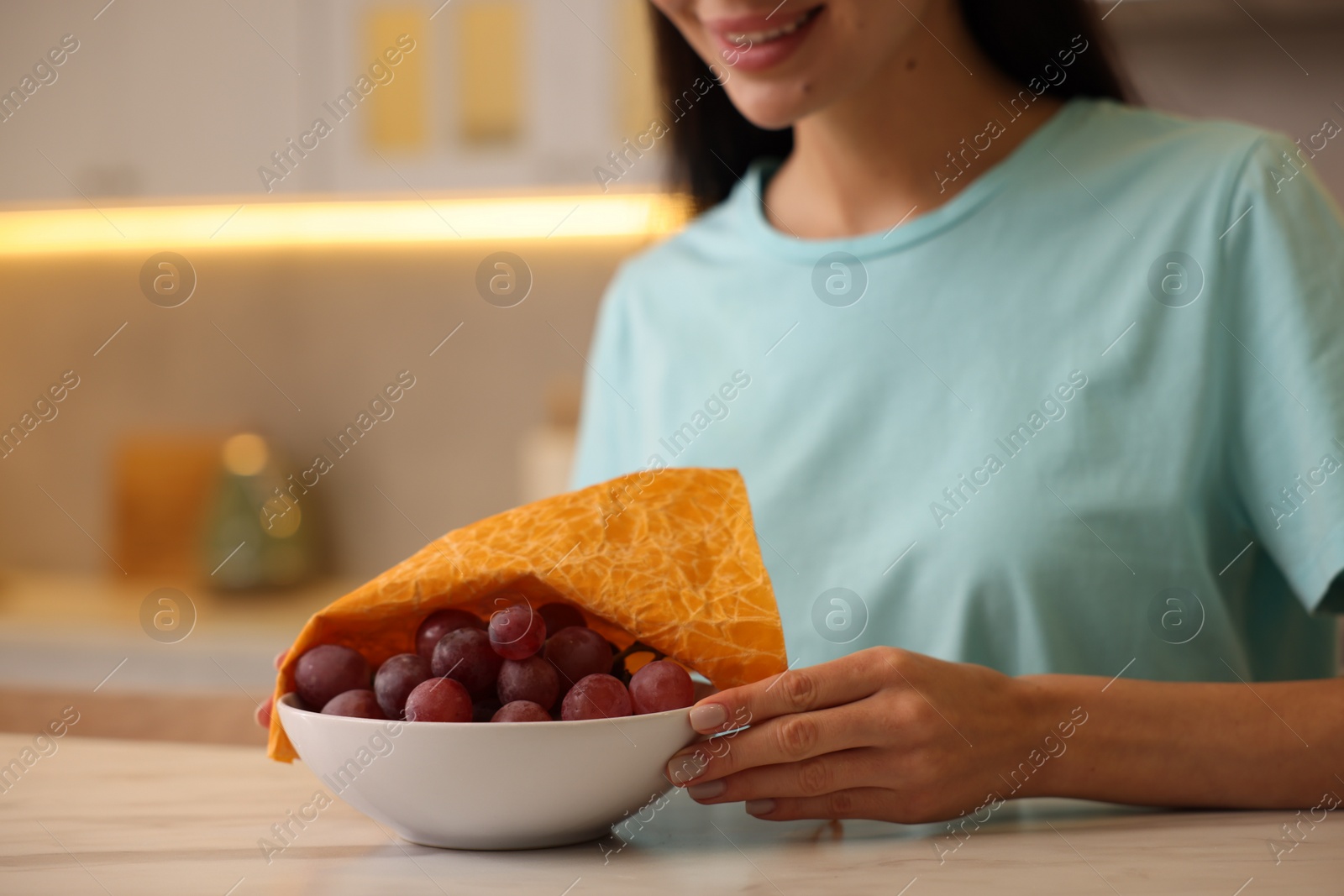 Photo of Woman packing bowl of fresh grapes into beeswax food wrap at table in kitchen, closeup