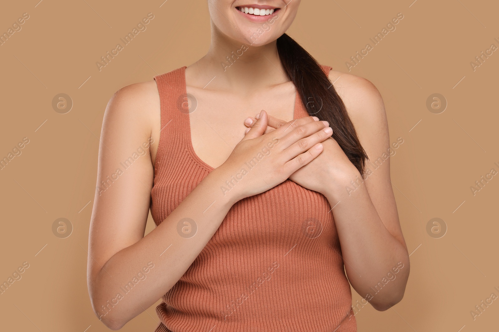 Photo of Thank you gesture. Grateful woman holding hands near heart on brown background, closeup