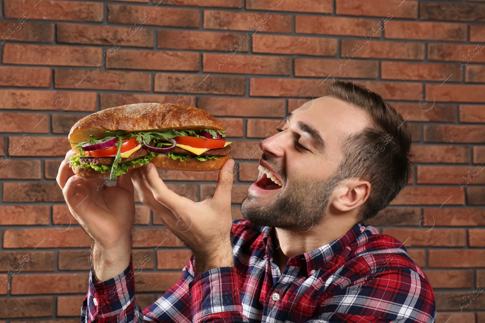 Photo of Young hungry man eating tasty sandwich on brick wall background