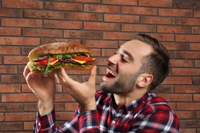 Young hungry man eating tasty sandwich on brick wall background
