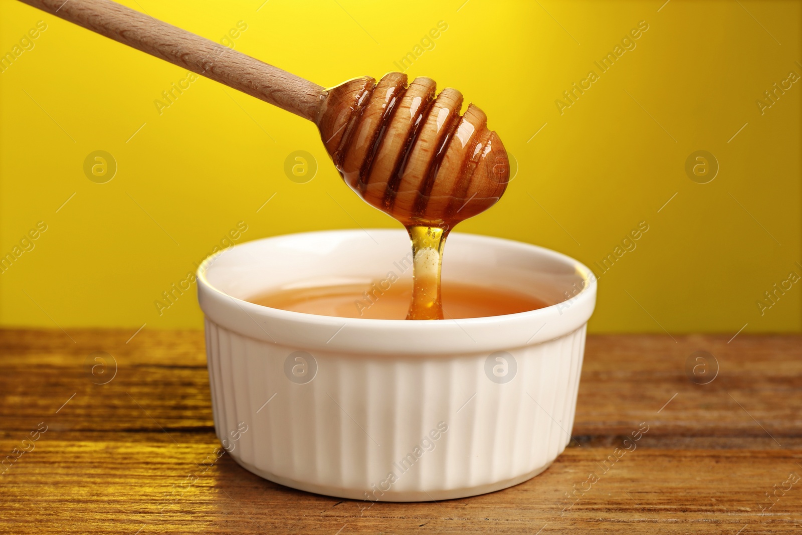 Photo of Pouring honey from dipper into bowl at wooden table against yellow background, closeup