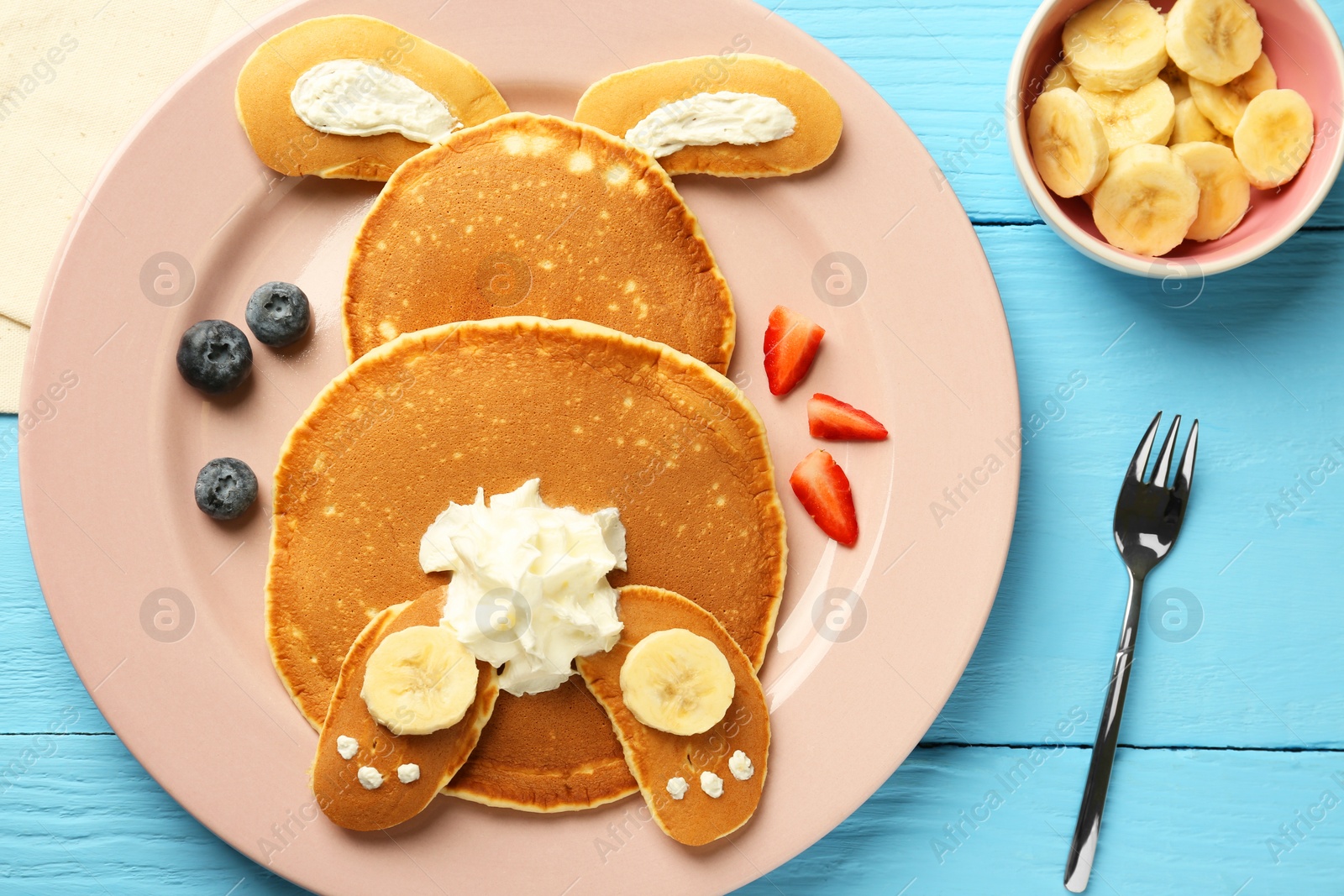 Photo of Creative serving for kids. Plate with cute bunny made of pancakes, berries, cream and banana on light blue wooden table, flat lay