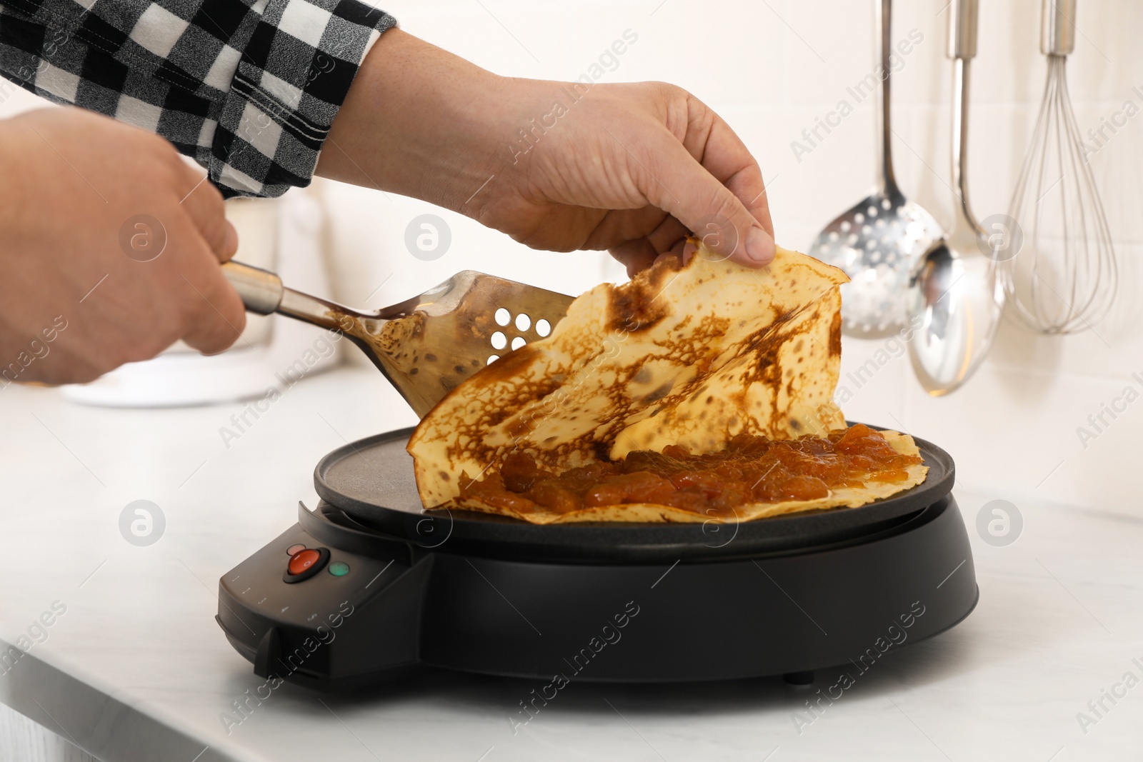 Photo of Man cooking delicious crepe with jam on electric pancake maker in kitchen, closeup