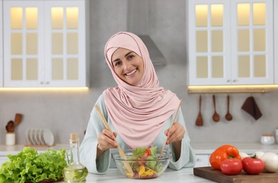 Photo of Muslim woman making delicious salad with vegetables at white table in kitchen