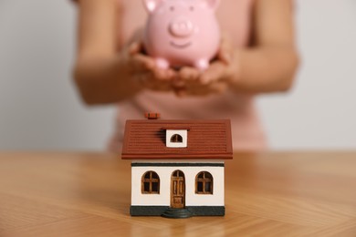 Photo of Woman holding piggy bank at wooden table, focus on little house model
