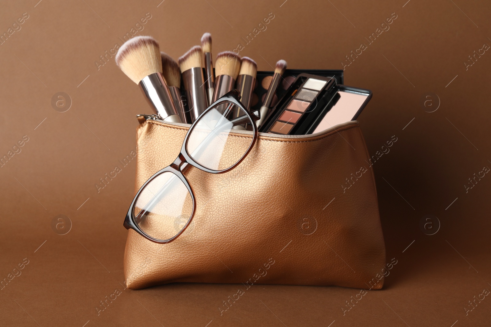 Photo of Cosmetic bag with makeup products and eyeglasses on brown background