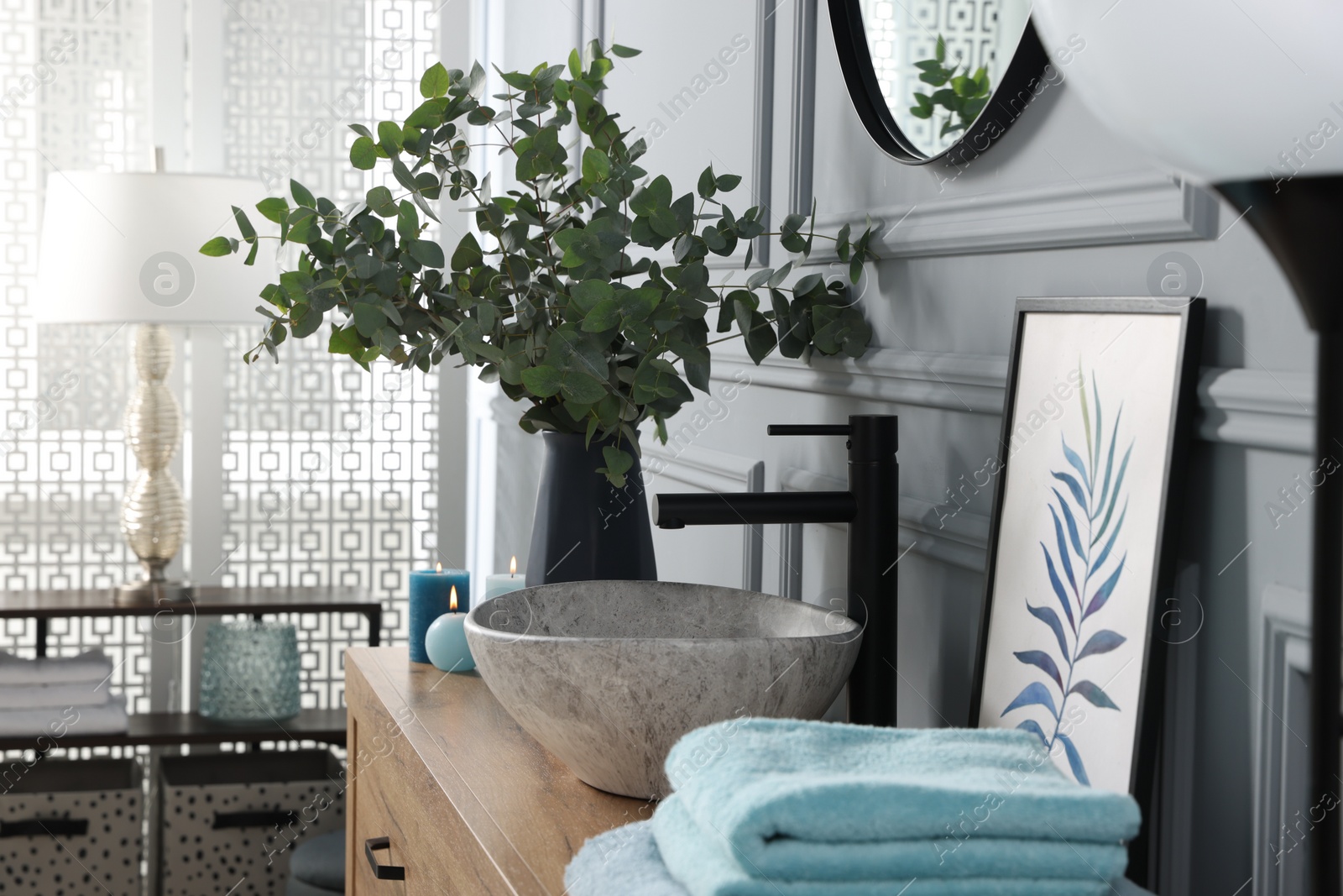 Photo of Eucalyptus branches near vessel sink, aroma candles and towels on bathroom vanity. Interior design