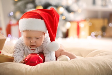 Little baby in Santa hat playing with Christmas balls indoors