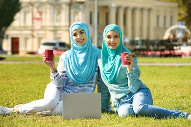 Muslim women in hijabs with laptop sitting on green lawn outdoors