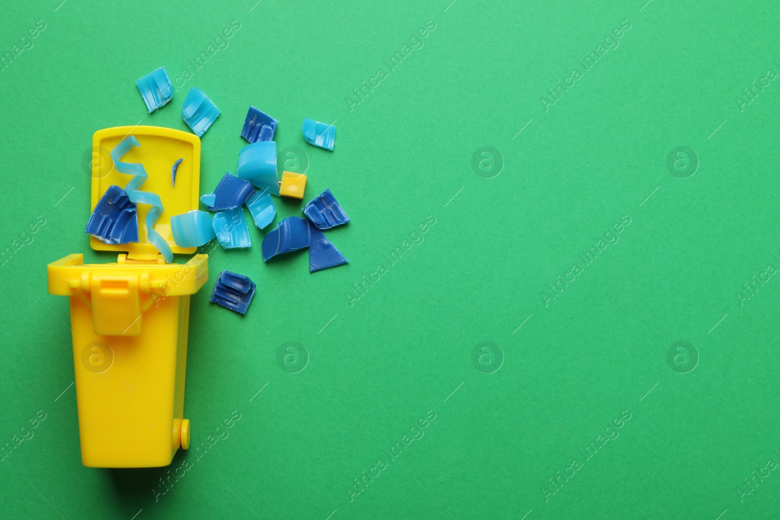 Photo of Mini recycling bin and broken plastic pieces on green background, flat lay. Space for text