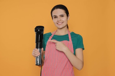 Photo of Beautiful young woman pointing on sous vide cooker against orange background