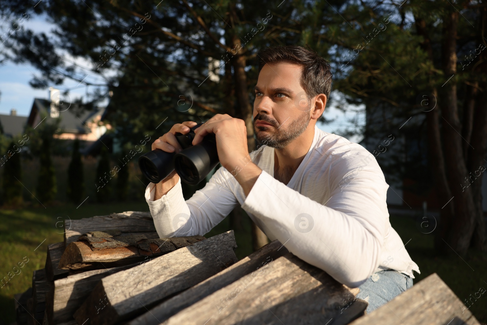 Photo of Concept of private life. Curious man with binoculars spying on neighbours over firewood outdoors