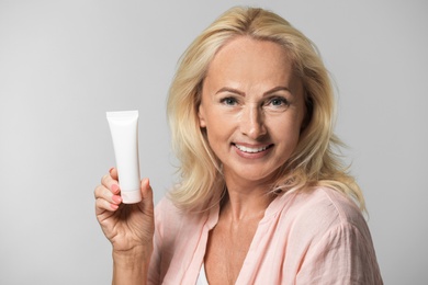 Photo of Portrait of beautiful mature woman with perfect skin holding tube of cream on grey background