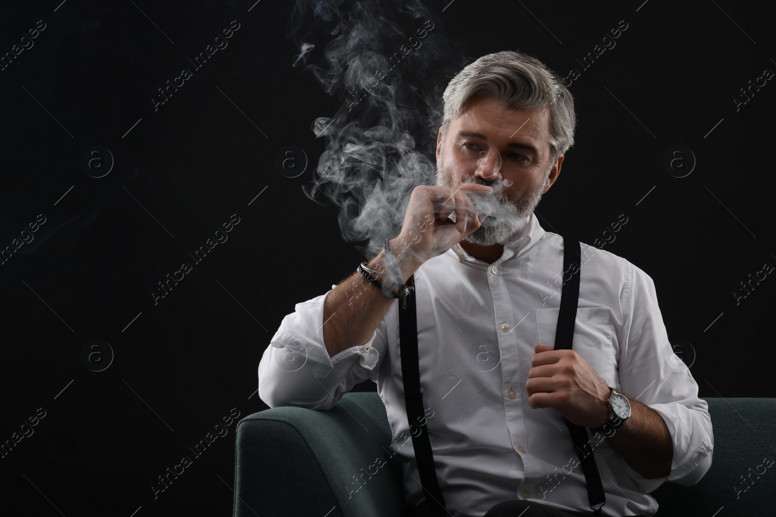 Photo of Bearded man smoking cigar on sofa against black background. Space for text