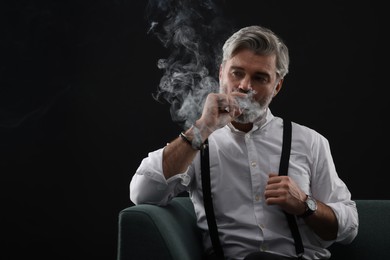 Photo of Bearded man smoking cigar on sofa against black background. Space for text