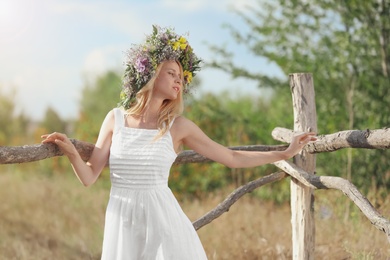 Young woman wearing wreath made of beautiful flowers near wooden fence on sunny day