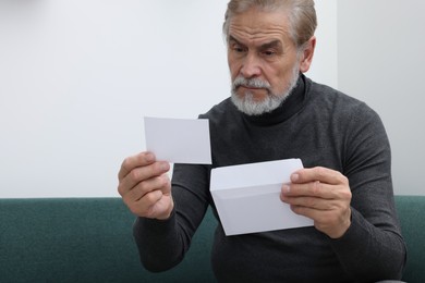 Upset senior man holding envelope and photo on sofa at home, space for text. Loneliness concept