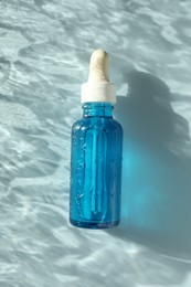 Wet bottle of cosmetic serum on light blue background, top view