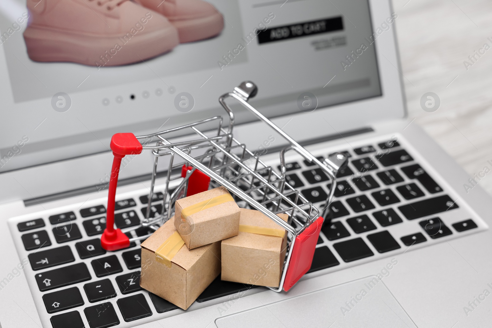 Photo of Internet store. Small cardboard boxes, shopping cart and laptop on table, closeup