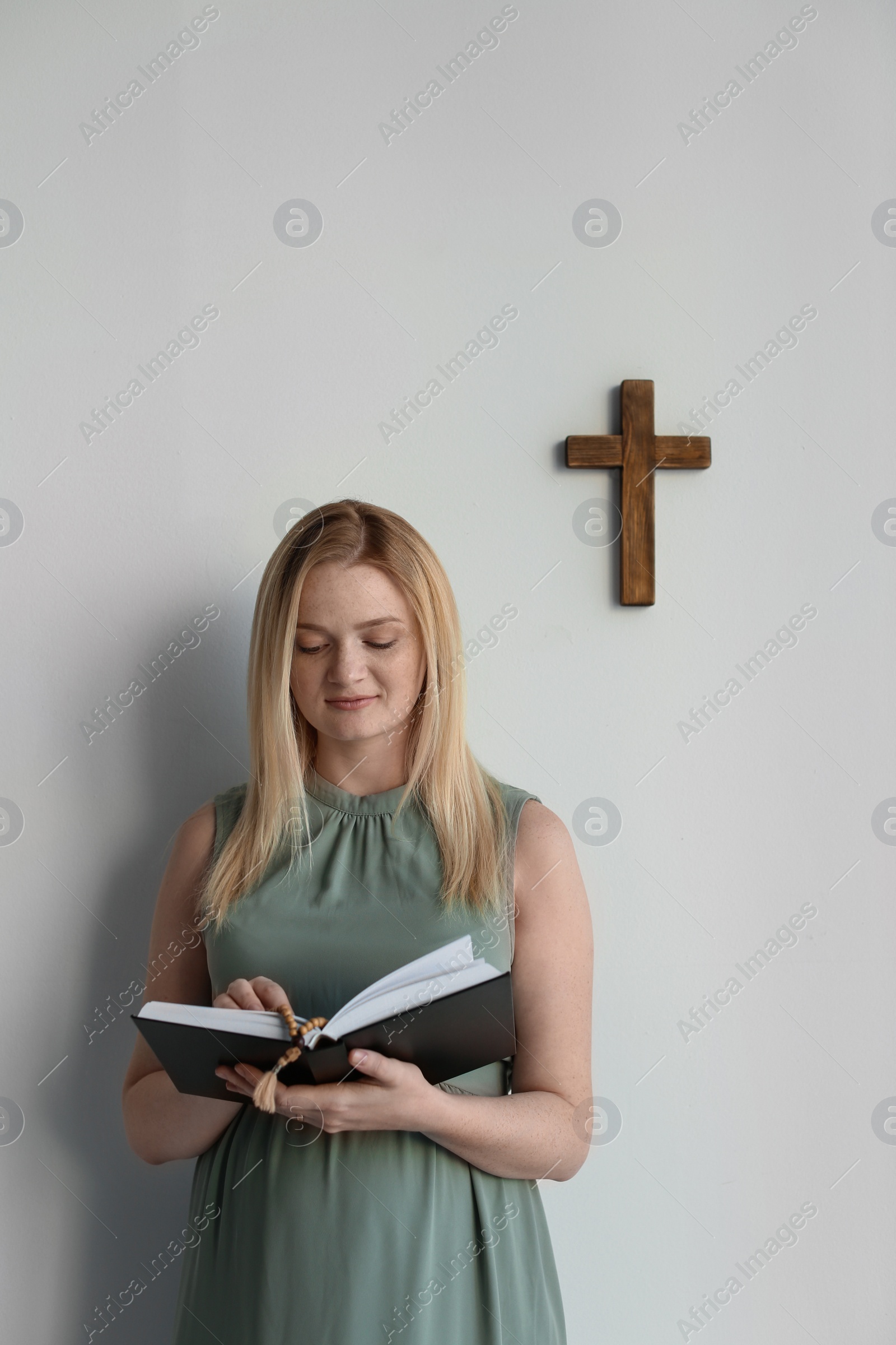 Photo of Young pregnant woman with Bible praying near light wall