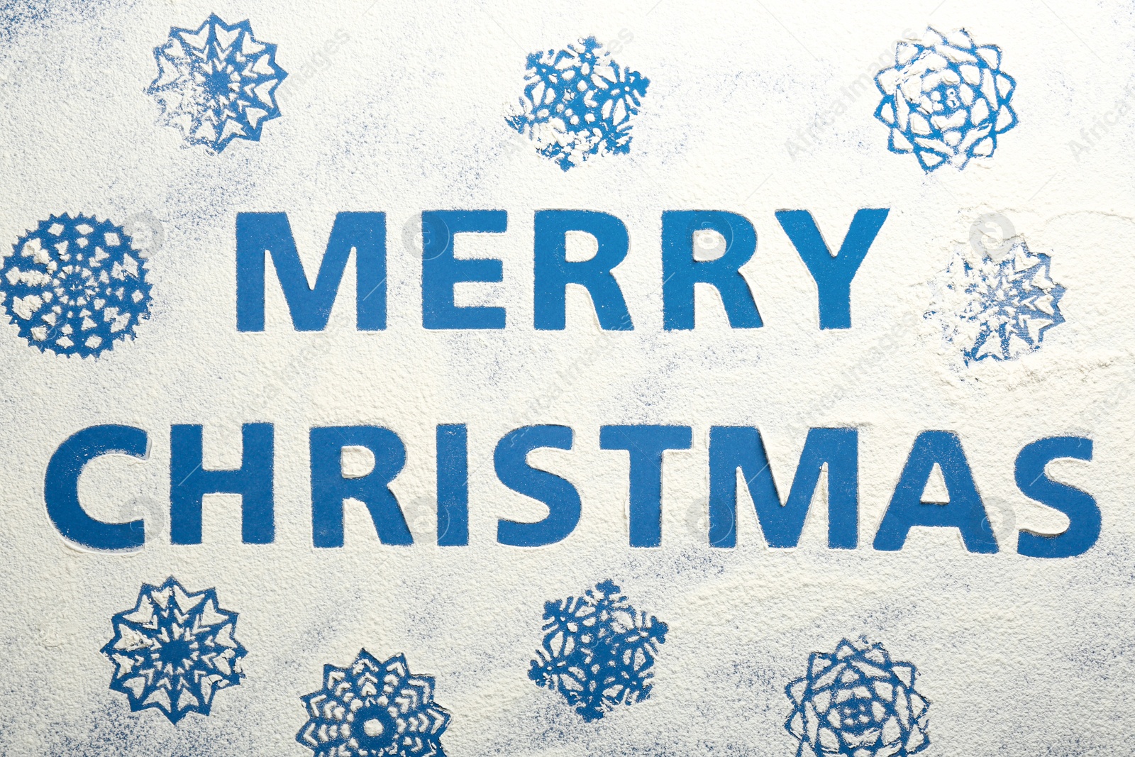 Photo of Words MERRY CHRISTMAS and snowflakes made of flour on blue background, top view