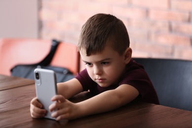 Photo of Sad little boy with mobile phone sitting at table indoors