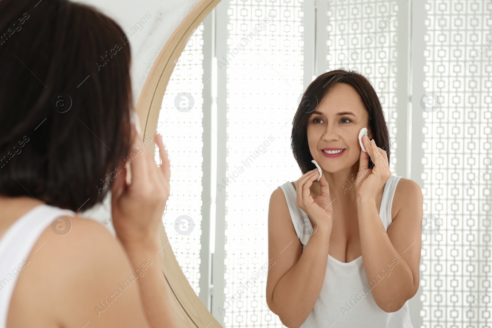 Photo of Mature woman cleaning face with cotton pads near mirror in bathroom