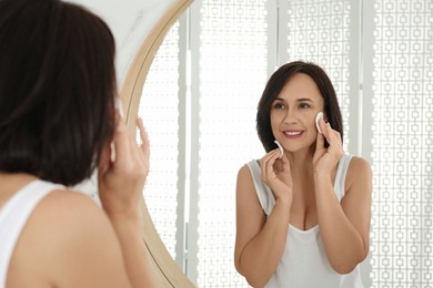 Mature woman cleaning face with cotton pads near mirror in bathroom