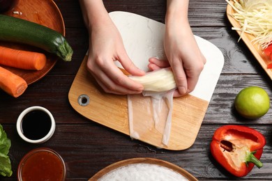 Making delicious spring rolls. Woman wrapping fresh vegetables into rice paper at wooden table, flat lay