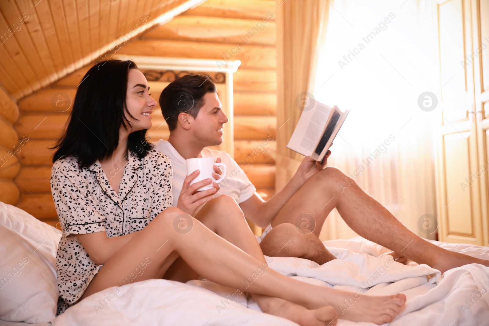 Photo of Happy couple enjoying lazy morning with drink and book in bedroom