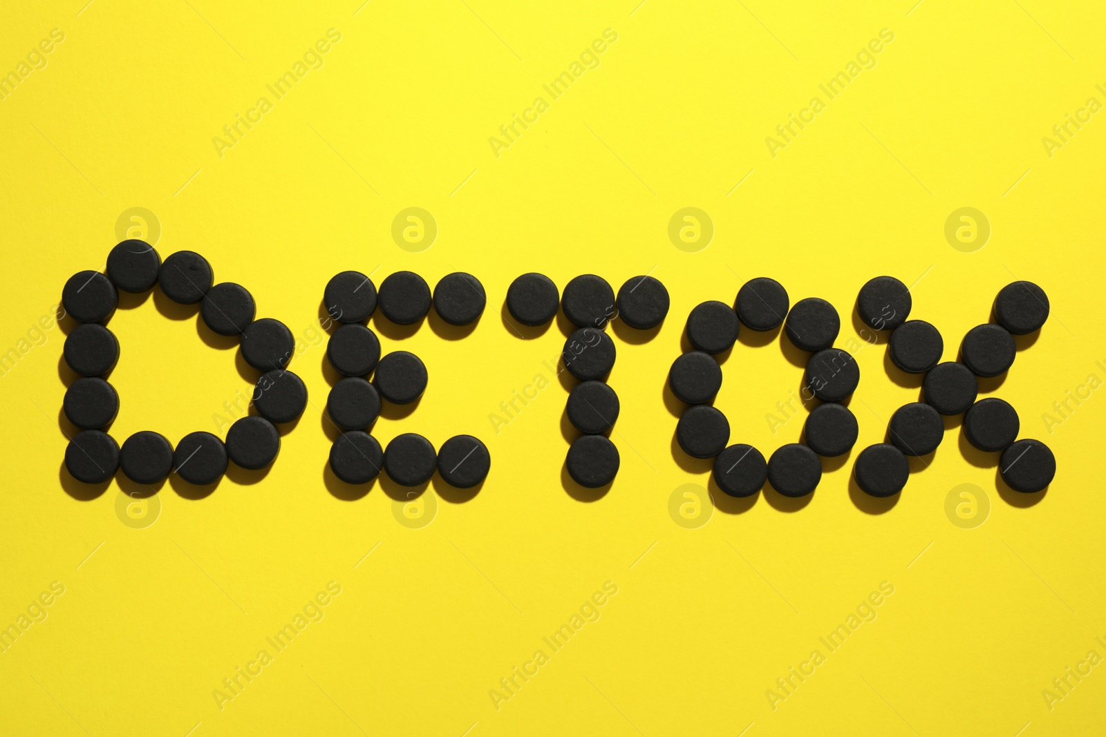 Photo of Word Detox made of activated charcoal pills on yellow background, flat lay. Potent sorbent
