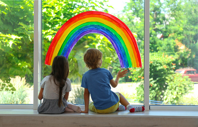 Photo of Little children sitting near rainbow painting on window indoors. Stay at home concept