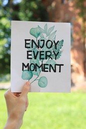 Photo of Woman holding card with phrase Enjoy Every Moment outdoors, closeup