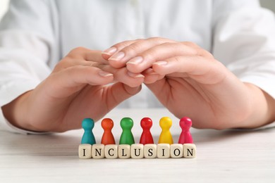 Photo of Woman protecting colorful pawns and wooden cubes with word Inclusion at white table, closeup