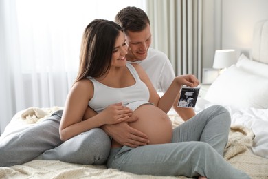 Young pregnant woman and her husband with ultrasound picture of baby in bedroom