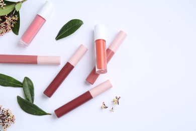 Different lip glosses, green leaves and flowers on white background, flat lay. Space for text