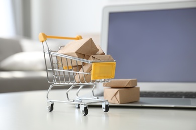 Photo of Internet shopping. Laptop and small cart with boxes on table indoors