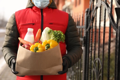 Photo of Courier in medical mask holding paper bag with groceries near house outdoors, closeup. Delivery service during quarantine due to Covid-19 outbreak