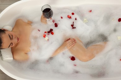 Photo of Woman with glass of wine taking bath in tub with foam and rose petals, top view