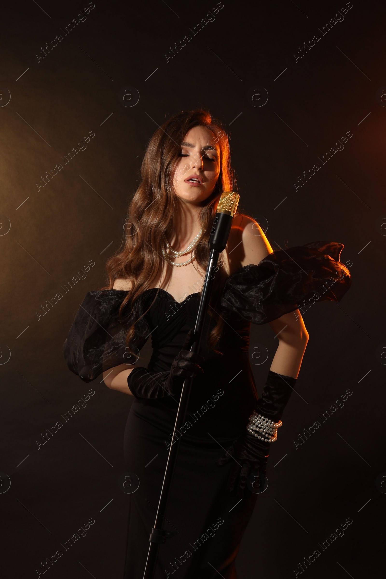 Photo of Beautiful young woman in stylish dress with microphone singing on dark background