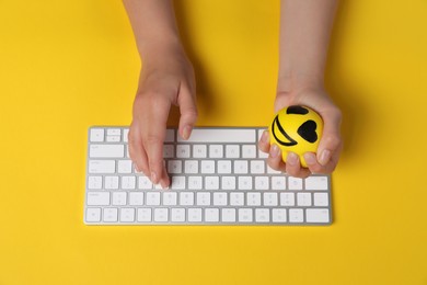 Photo of Woman squeezing antistress ball while typing on keyboard against yellow background, top view