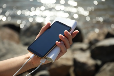 Photo of Woman charging smartphone with power bank on rocky mountain near river, closeup